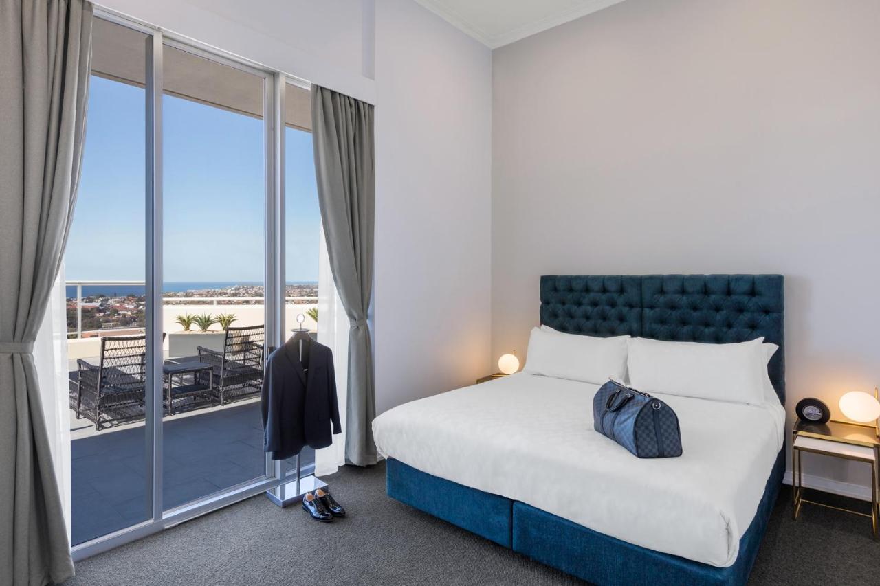 เมอริตัน สวีท บอนดี จังค์ชั่น Hotel ซิดนีย์ ภายนอก รูปภาพ The photo shows a stylish bedroom featuring a large bed with a tufted blue headboard. There's a neatly organized outfit consisting of a suit hanging on a stand, along with shoes placed on the floor. The room has large windows that provide a view of t