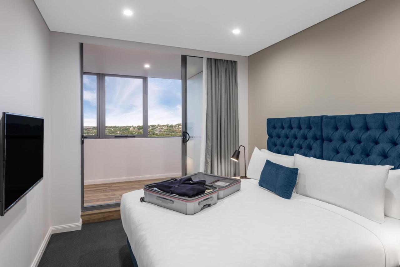 เมอริตัน สวีท บอนดี จังค์ชั่น Hotel ซิดนีย์ ภายนอก รูปภาพ The photo shows a modern hotel room featuring a double bed with a tufted blue headboard. There is a neatly arranged suitcase placed open on the bed, indicating that someone may be unpacking. The room has light-colored walls and a window allowing natu