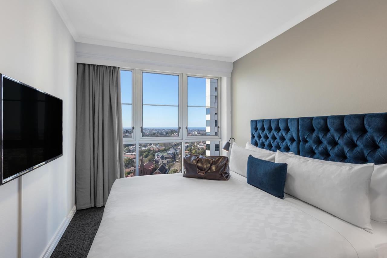 เมอริตัน สวีท บอนดี จังค์ชั่น Hotel ซิดนีย์ ภายนอก รูปภาพ The photo shows a modern hotel room featuring a large bed with a tufted blue headboard. Beside the bed, there is a stylish bag placed on the bed. The room has large windows that provide a view of the outside, allowing natural light to fill the space.