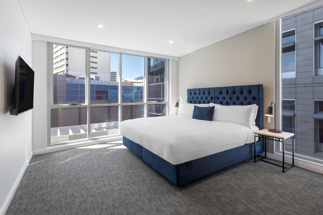 เมอริตัน สวีท บอนดี จังค์ชั่น Hotel ซิดนีย์ ภายนอก รูปภาพ The image shows a modern hotel room with a large, comfortable bed featuring a blue upholstered headboard. The room has large windows that allow natural light to enter, providing a good view of the surrounding buildings. There is a small bedside table