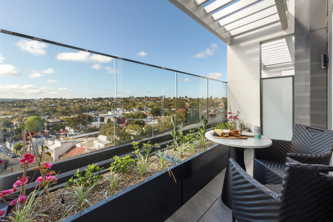 เมอริตัน สวีท บอนดี จังค์ชั่น Hotel ซิดนีย์ ภายนอก รูปภาพ The photo shows a modern balcony with a glass railing, enhancing the view of the surrounding area, which includes various rooftops and greenery in the distance. There is a small round table set with a decorative floral arrangement and some items, lik