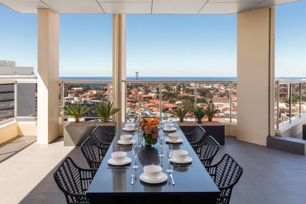 เมอริตัน สวีท บอนดี จังค์ชั่น Hotel ซิดนีย์ ภายนอก รูปภาพ The photo shows a modern outdoor dining area situated on a balcony or terrace with a stunning view. There is a long rectangular table set with plates, glasses, and cutlery, ready for a meal. The table is surrounded by sleek, black wicker chairs. In t