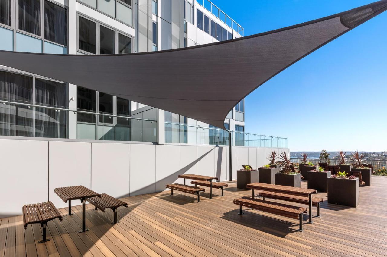 เมอริตัน สวีท บอนดี จังค์ชั่น Hotel ซิดนีย์ ภายนอก รูปภาพ The photo shows a modern outdoor terrace area on a building. It features several wooden benches arranged on a wooden deck. There are large planters with greenery, adding some decorative elements. A large triangular gray shade sail is positioned above