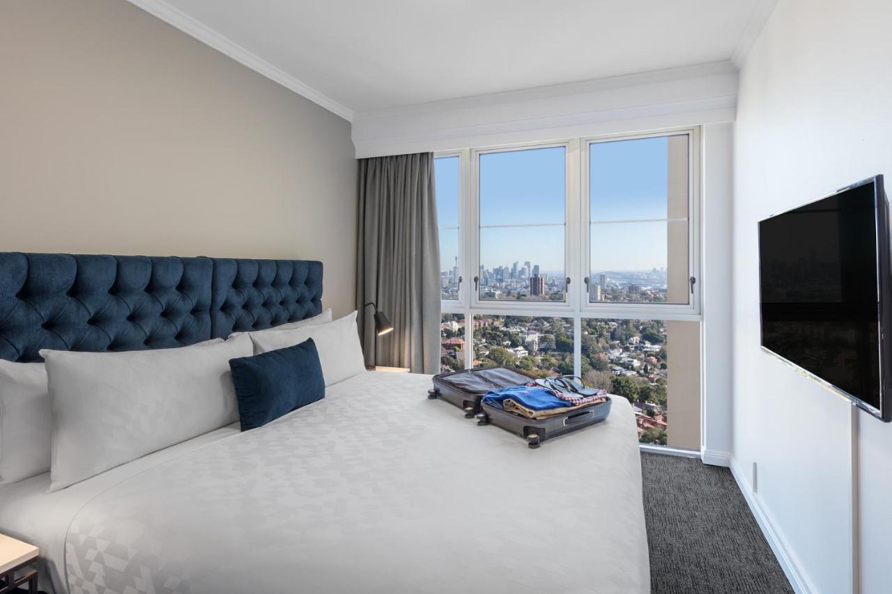 เมอริตัน สวีท บอนดี จังค์ชั่น Hotel ซิดนีย์ ภายนอก รูปภาพ The photo shows a modern hotel bedroom featuring a king-sized bed with a blue tufted headboard. The bed is neatly made with white linens and a decorative blue pillow. There is a large window that offers a view of the city skyline, allowing natural li