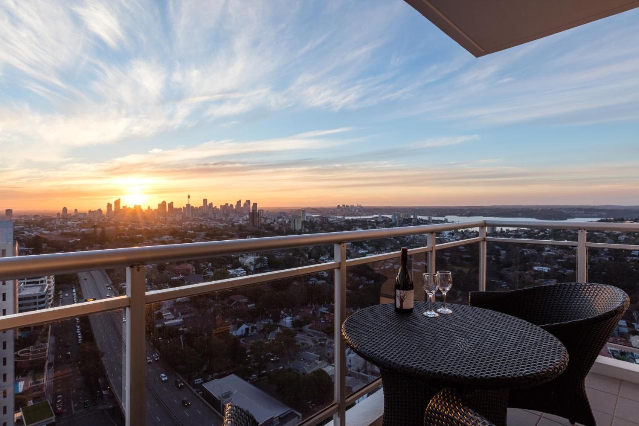 เมอริตัน สวีท บอนดี จังค์ชั่น Hotel ซิดนีย์ ภายนอก รูปภาพ The image shows a scenic view from a balcony or terrace overlooking a cityscape during sunset. In the foreground, there is a small round table with two glasses and a bottle of wine, suggesting a romantic or relaxing setting. The sky is filled with vi