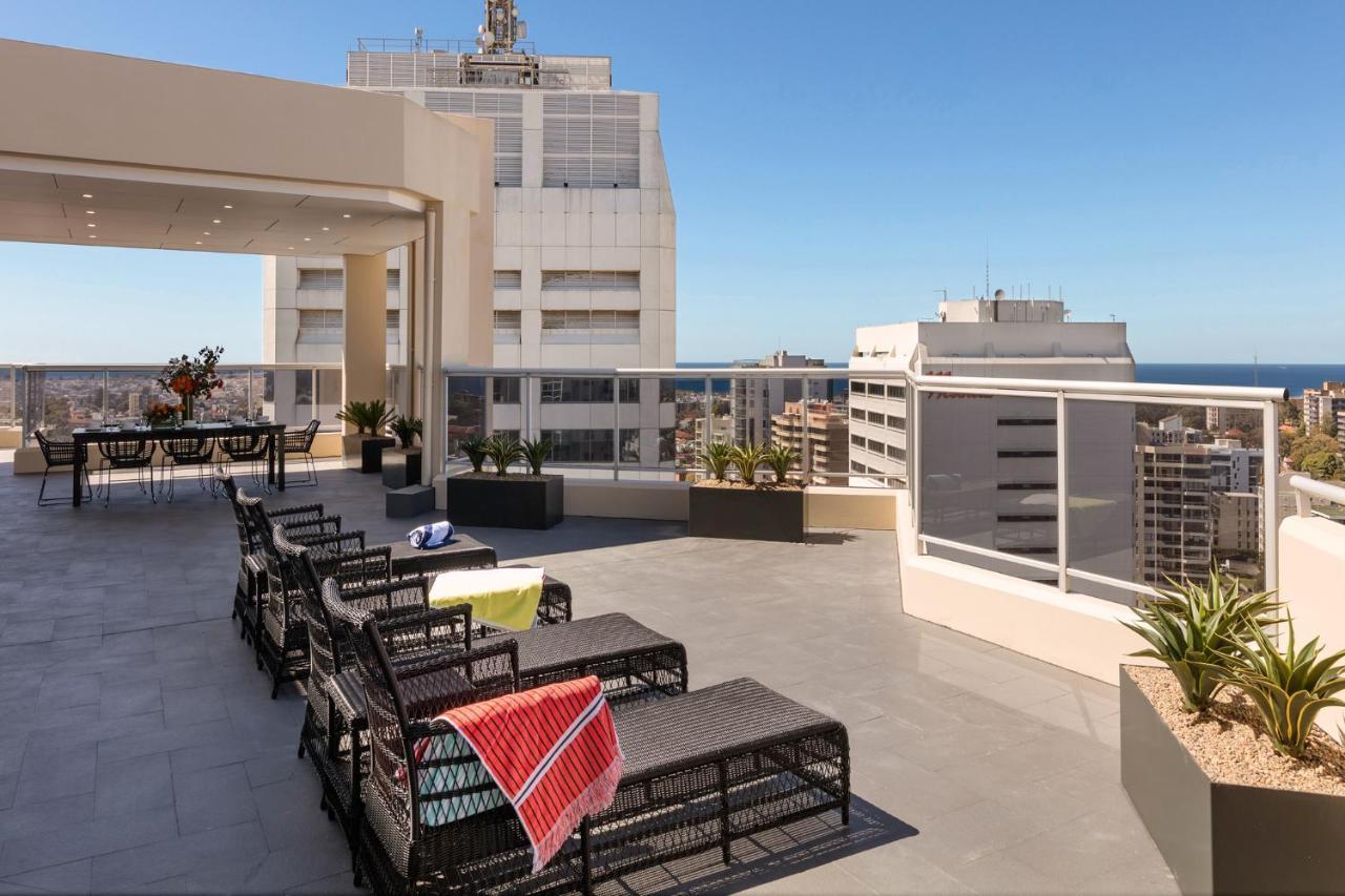 เมอริตัน สวีท บอนดี จังค์ชั่น Hotel ซิดนีย์ ภายนอก รูปภาพ The photo shows a rooftop terrace with a modern design. There are several outdoor seating arrangements featuring black wicker lounge chairs. Some of the chairs have colorful blankets draped over them. The terrace is surrounded by a railing and has po