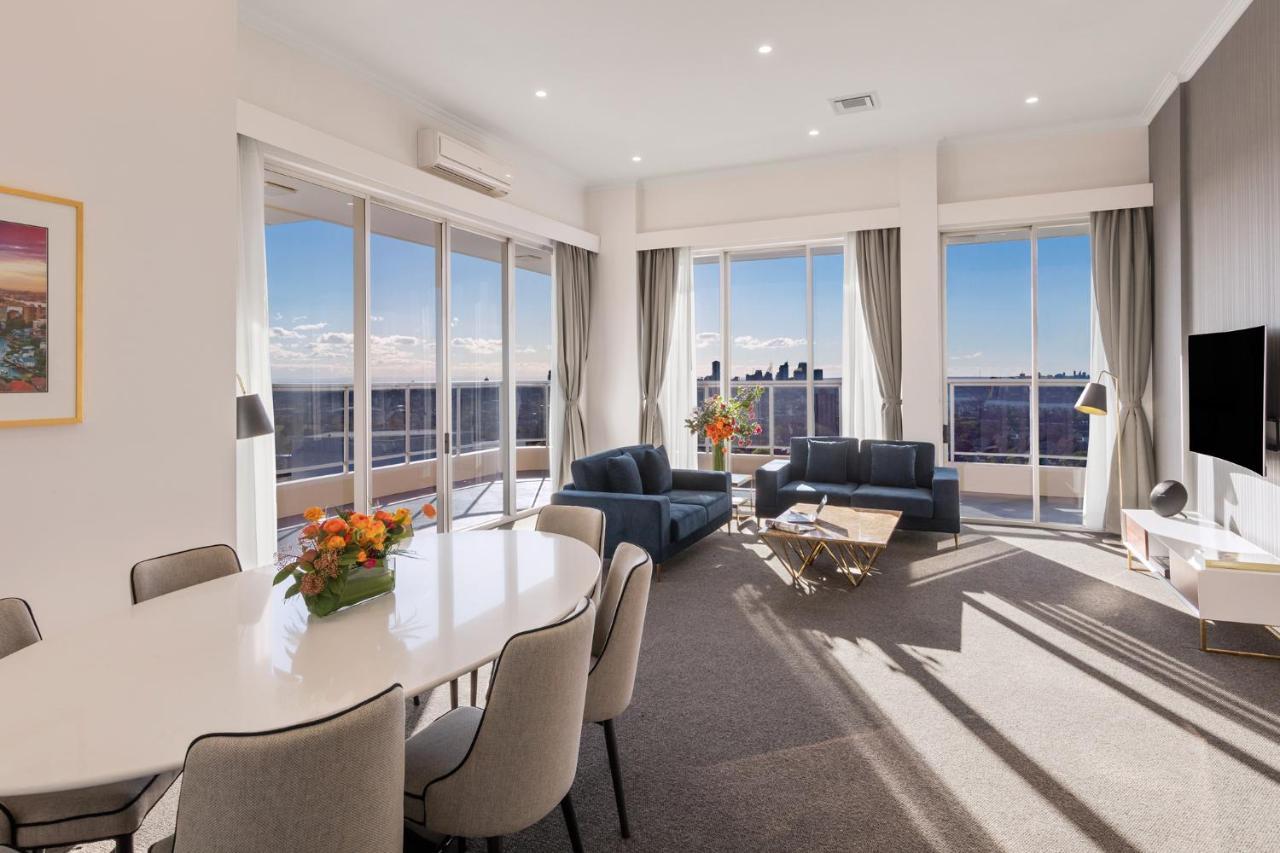 เมอริตัน สวีท บอนดี จังค์ชั่น Hotel ซิดนีย์ ภายนอก รูปภาพ The photo shows a modern and stylish living space with large windows allowing natural light to fill the room. In the foreground, there is a round dining table with several chairs around it, adorned with a floral centerpiece. The seating area features