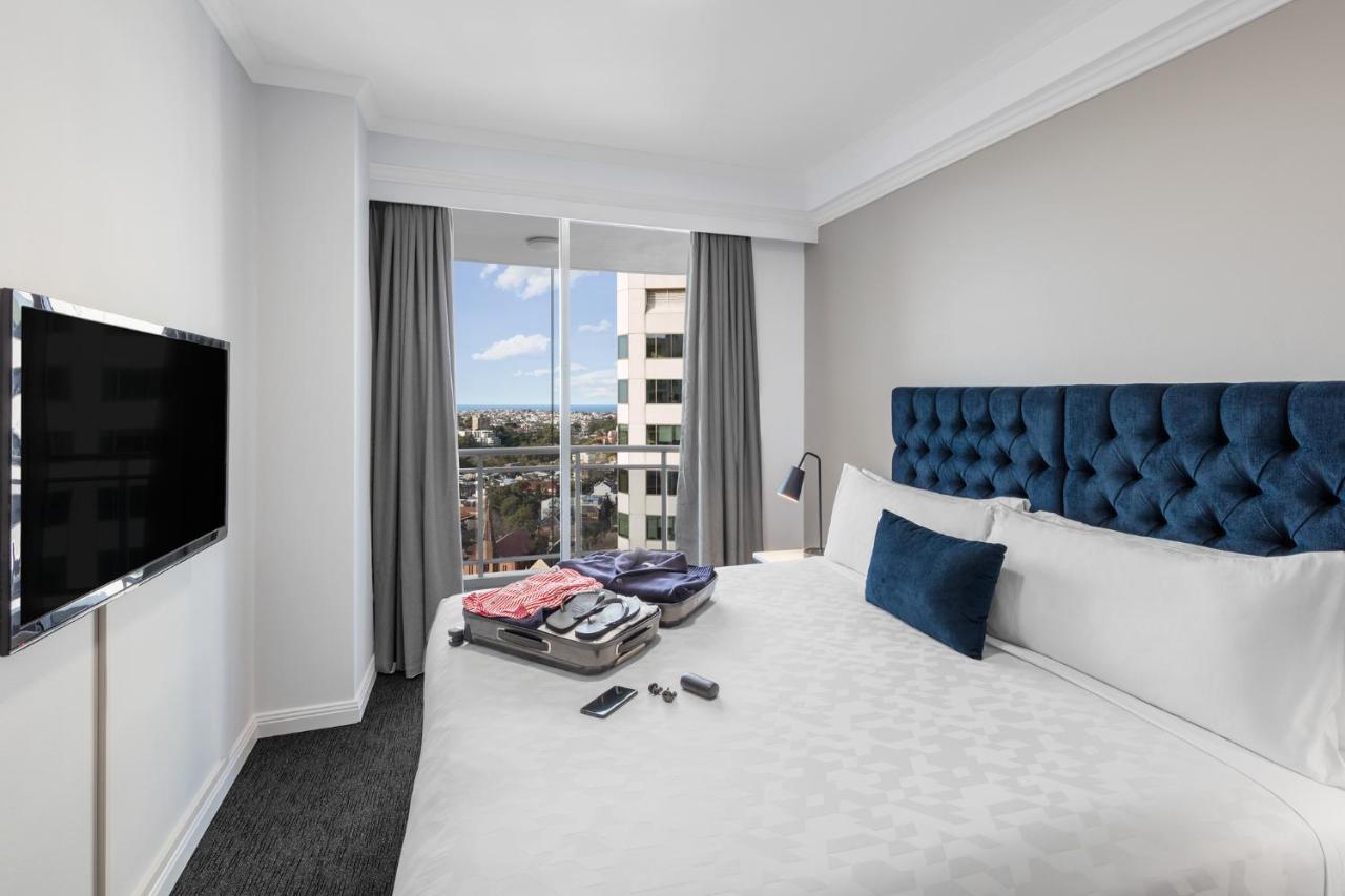 เมอริตัน สวีท บอนดี จังค์ชั่น Hotel ซิดนีย์ ภายนอก รูปภาพ The photo shows a modern hotel room featuring a neatly made bed with a blue tufted headboard and a white duvet. There's a small decorative pillow on the bed, and a piece of luggage is placed at the foot with clothes partially unpacked. A television i
