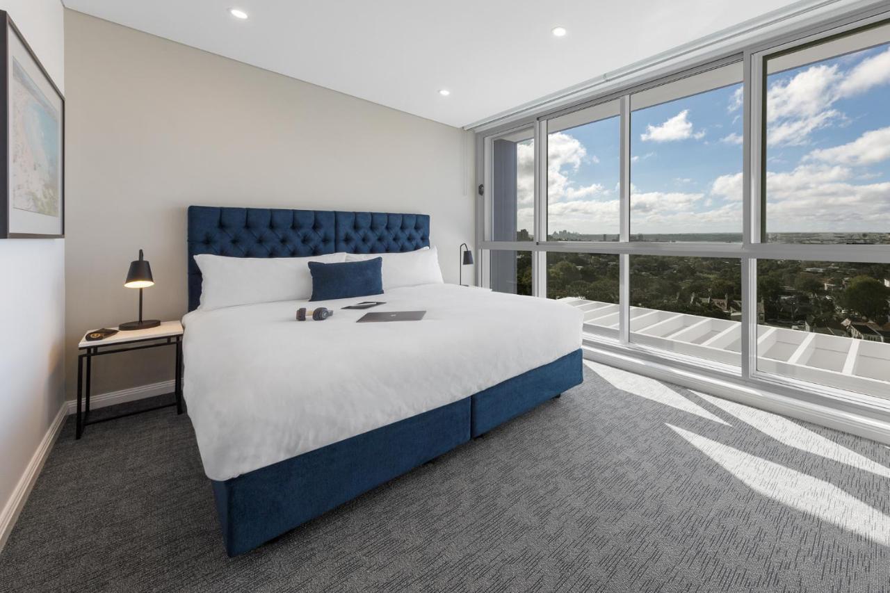 เมอริตัน สวีท บอนดี จังค์ชั่น Hotel ซิดนีย์ ภายนอก รูปภาพ The photo shows a modern and stylish hotel room. It features a comfortable queen-size bed with a blue tufted headboard. The bed is neatly made with white linens and pillows. Next to the bed, there is a small bedside table with a desk lamp. 

On the f