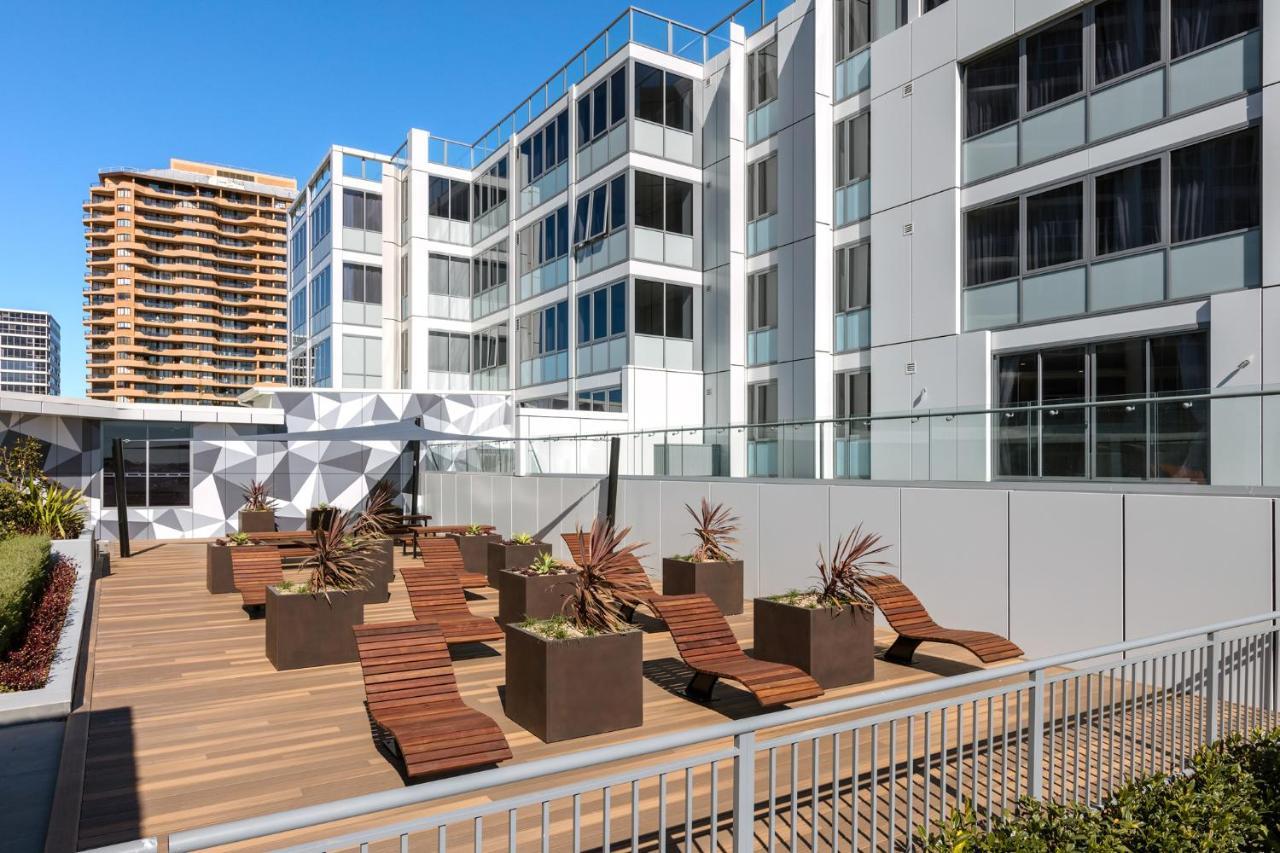 เมอริตัน สวีท บอนดี จังค์ชั่น Hotel ซิดนีย์ ภายนอก รูปภาพ The photo shows a modern outdoor space featuring a wooden deck. There are several lounge chairs arranged around large planter boxes, which contain plants. In the background, there are contemporary residential buildings with large windows, and a geome