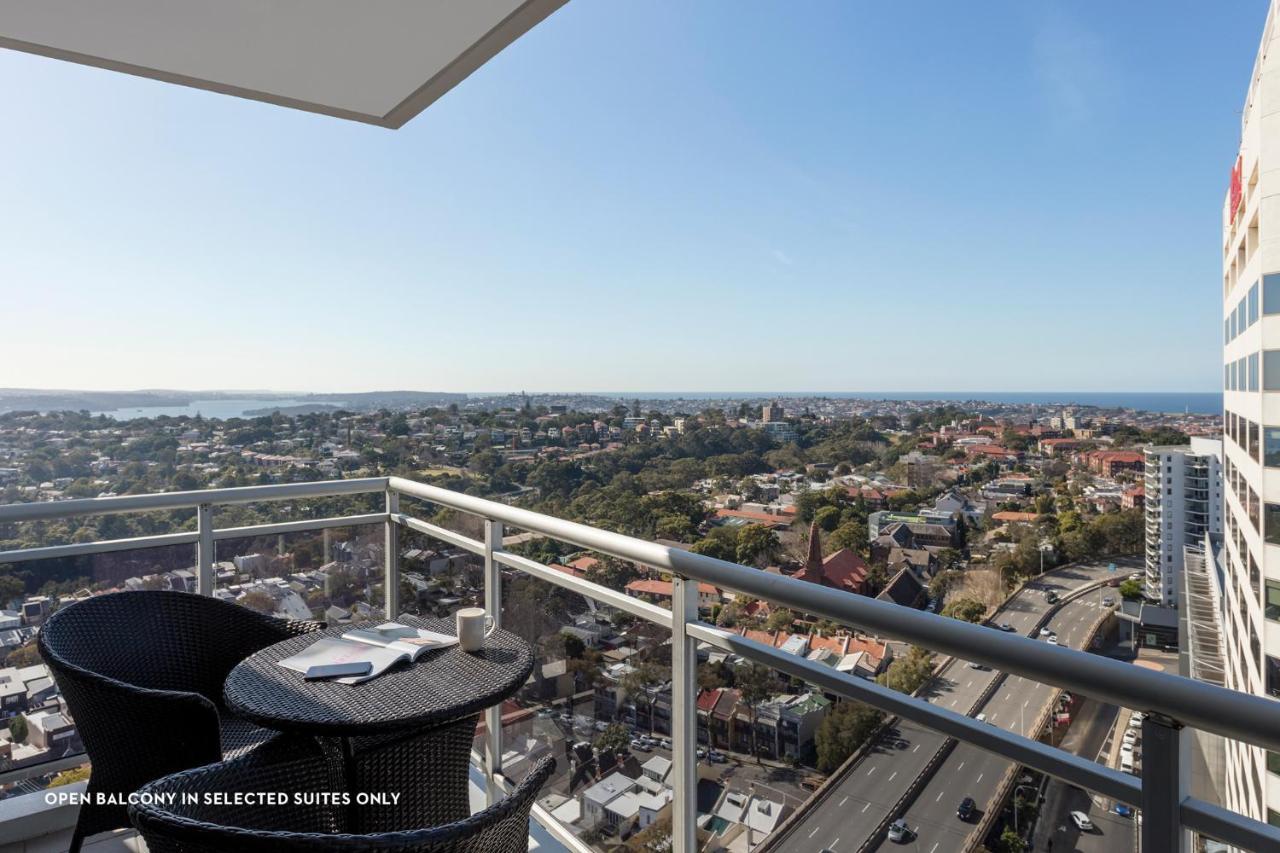 เมอริตัน สวีท บอนดี จังค์ชั่น Hotel ซิดนีย์ ภายนอก รูปภาพ The photo shows a balcony with a view over a cityscape that includes buildings and greenery, extending towards the horizon where the sea is visible. The balcony features a small round table and two black chairs, suggesting a space for relaxation or d