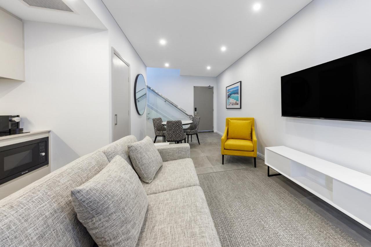 เมอริตัน สวีท บอนดี จังค์ชั่น Hotel ซิดนีย์ ภายนอก รูปภาพ The photo shows a modern and stylish living space. It features a light-colored sofa with cushions, positioned to face a wall-mounted flat-screen TV. There's a yellow accent chair nearby that adds a pop of color to the room. The décor is minimalistic,