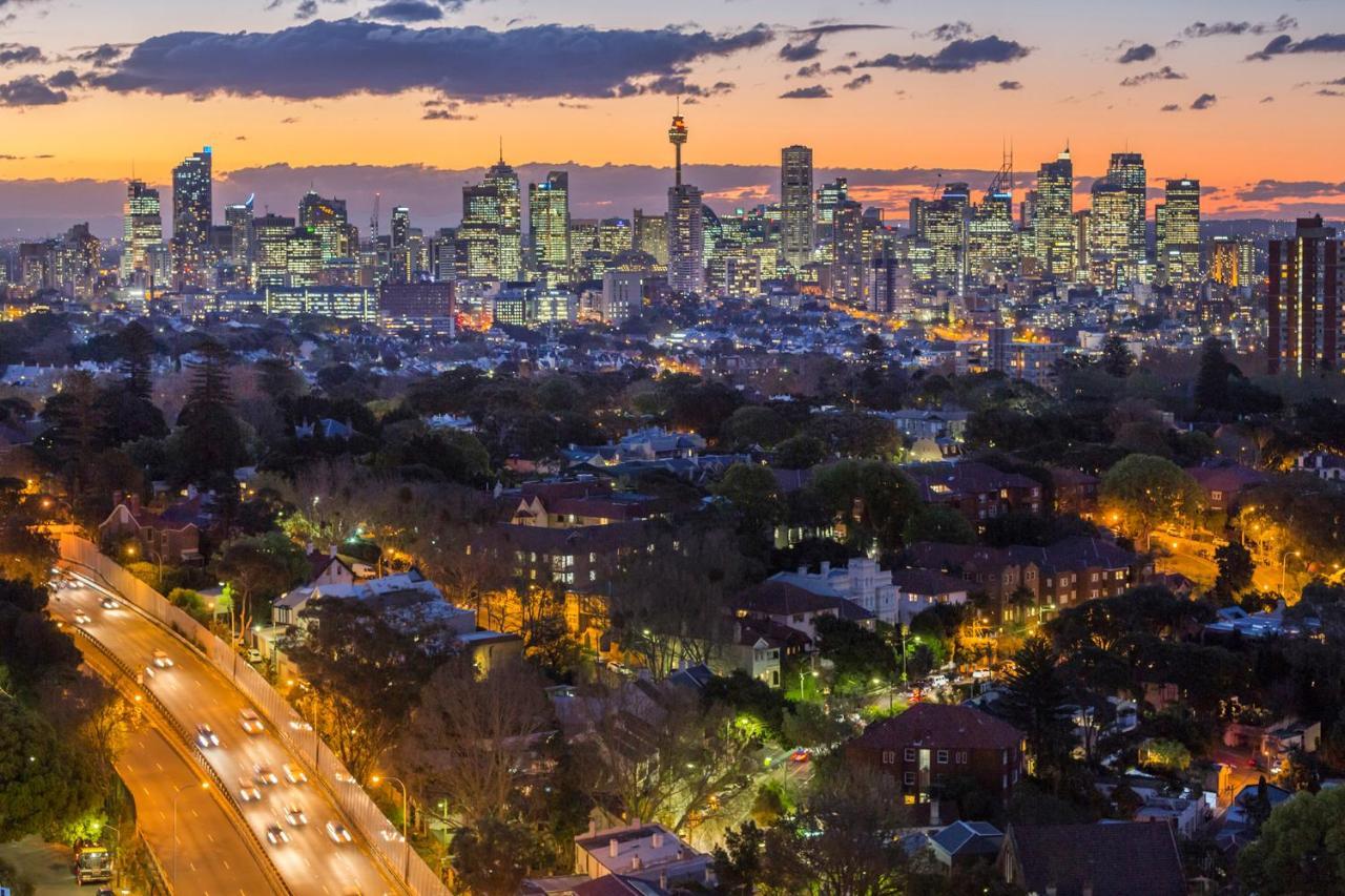 เมอริตัน สวีท บอนดี จังค์ชั่น Hotel ซิดนีย์ ภายนอก รูปภาพ The photo captures a stunning city skyline at dusk, featuring towering skyscrapers adorned with lights against a colorful sunset backdrop. In the foreground, a green area with trees and houses is visible, while a road with cars can be seen winding th