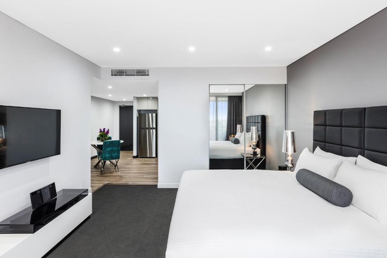 เมอริตัน สวีท บอนดี จังค์ชั่น Hotel ซิดนีย์ ภายนอก รูปภาพ The photo shows a modern hotel room. It features a large bed with a white covering and gray accent pillows. To the left, there's a flat-screen TV mounted on the wall above a sleek white cabinet. The flooring is a light wood tone, and a large mirror i