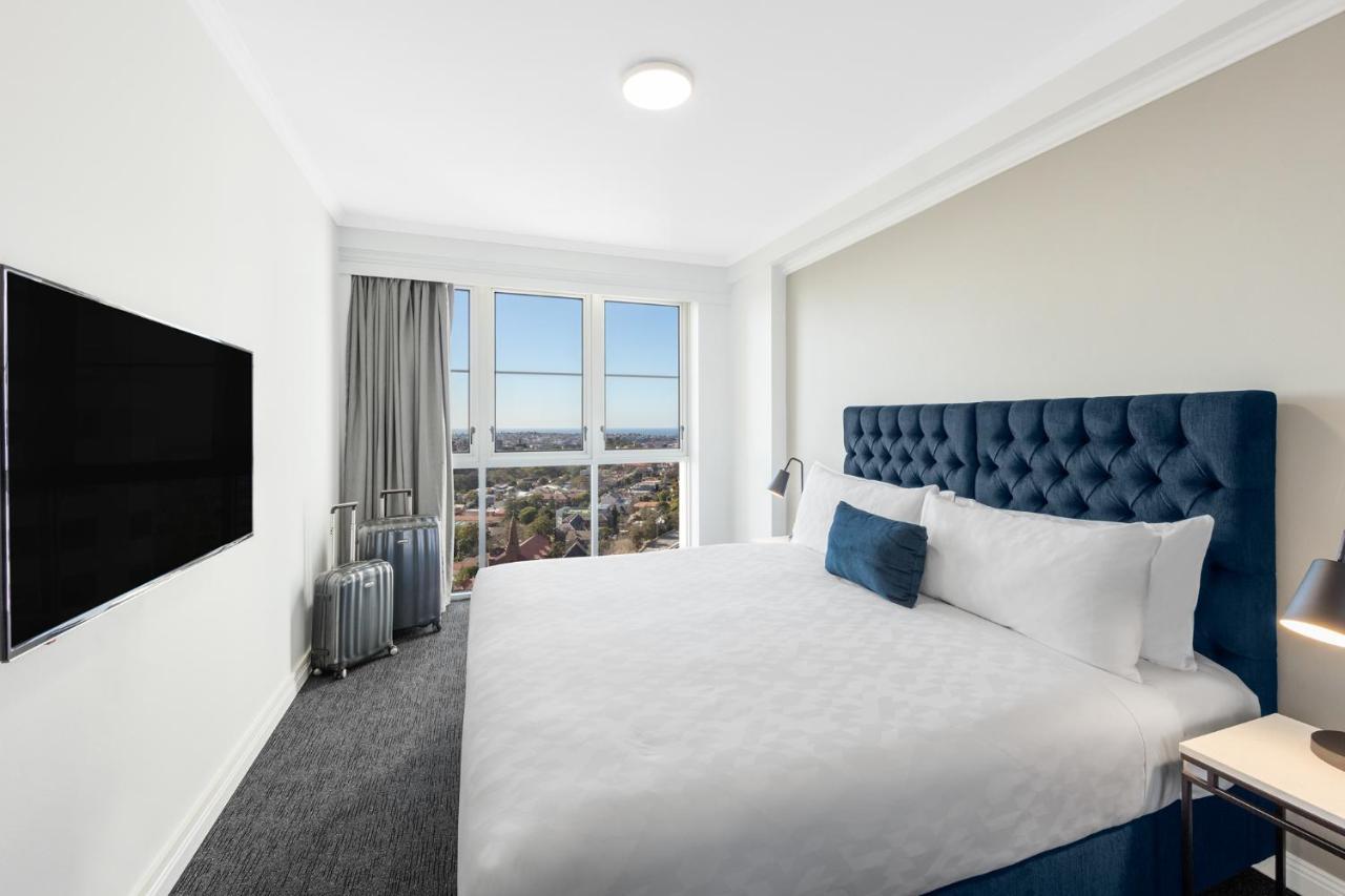 เมอริตัน สวีท บอนดี จังค์ชั่น Hotel ซิดนีย์ ภายนอก รูปภาพ The photo shows a modern hotel room with a large bed featuring a tufted blue headboard. There is a white duvet and a decorative pillow on the bed. A flat-screen television is mounted on the wall opposite the bed. Large windows are covered with gray c