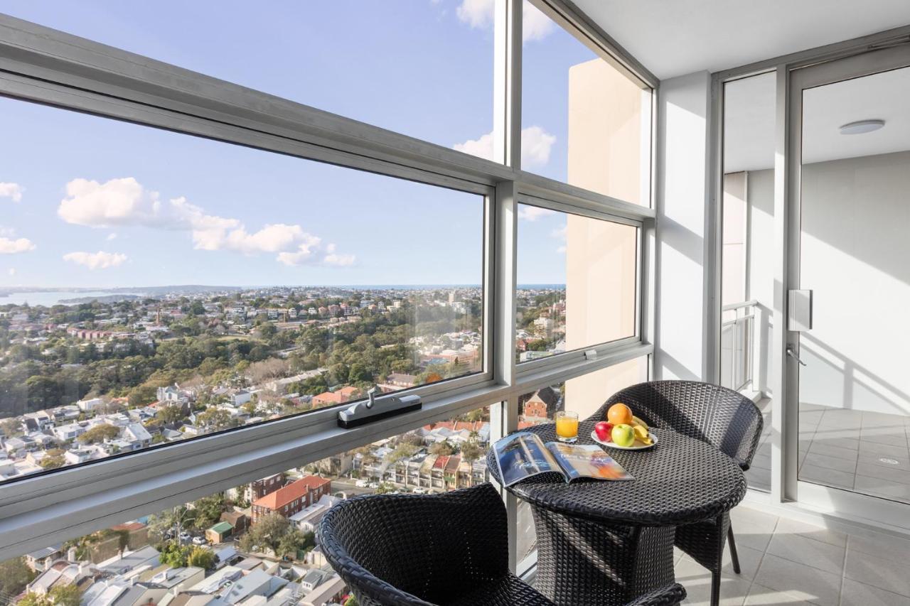 เมอริตัน สวีท บอนดี จังค์ชั่น Hotel ซิดนีย์ ภายนอก รูปภาพ The photo shows a modern balcony with large glass windows that offer a panoramic view of a sprawling urban landscape. There is a small round table made of dark wicker, accompanied by two matching chairs. On the table, there is a bowl of fruit and a m